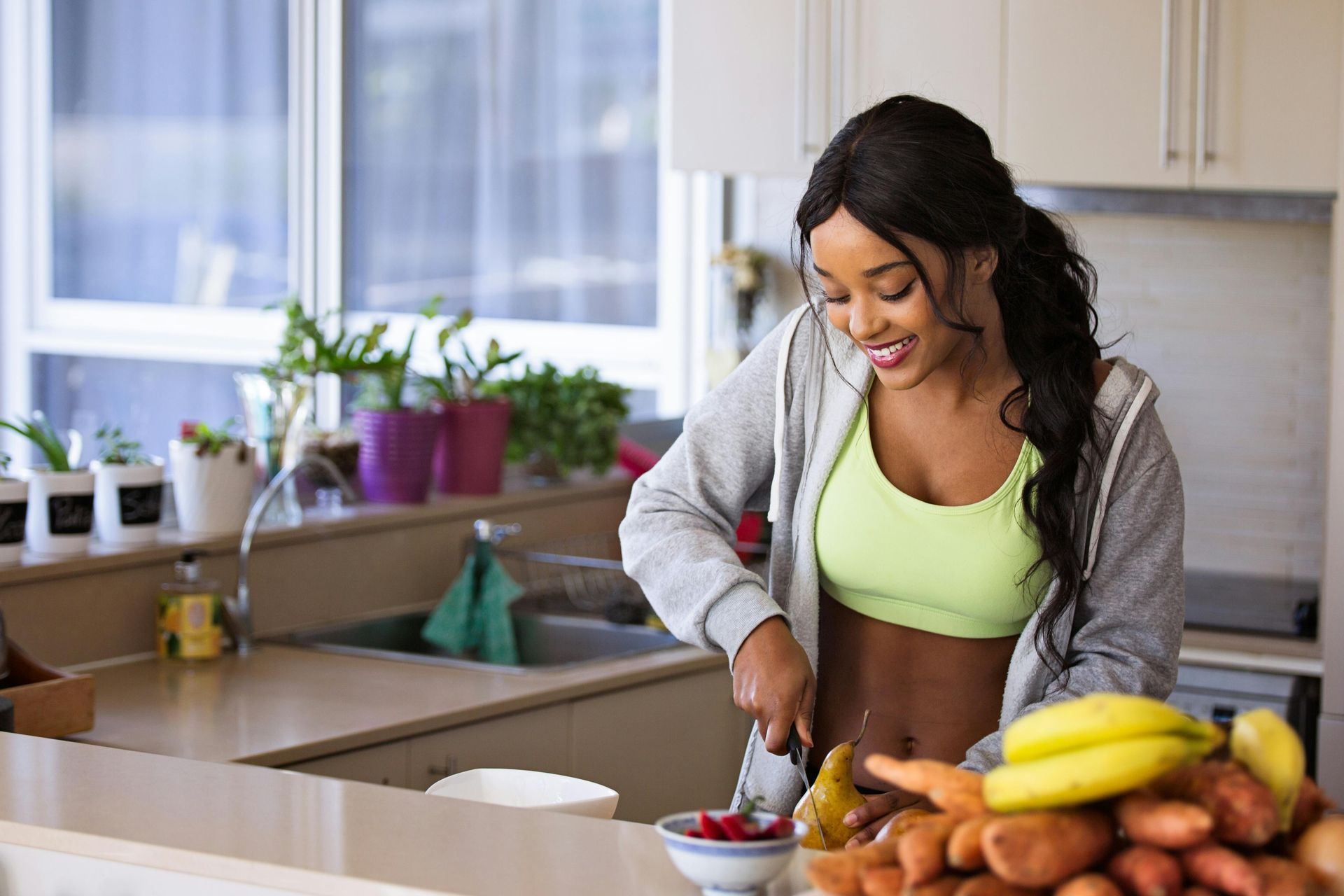 Woman in activewear, smiling, cutting fruit in a bright kitchen.