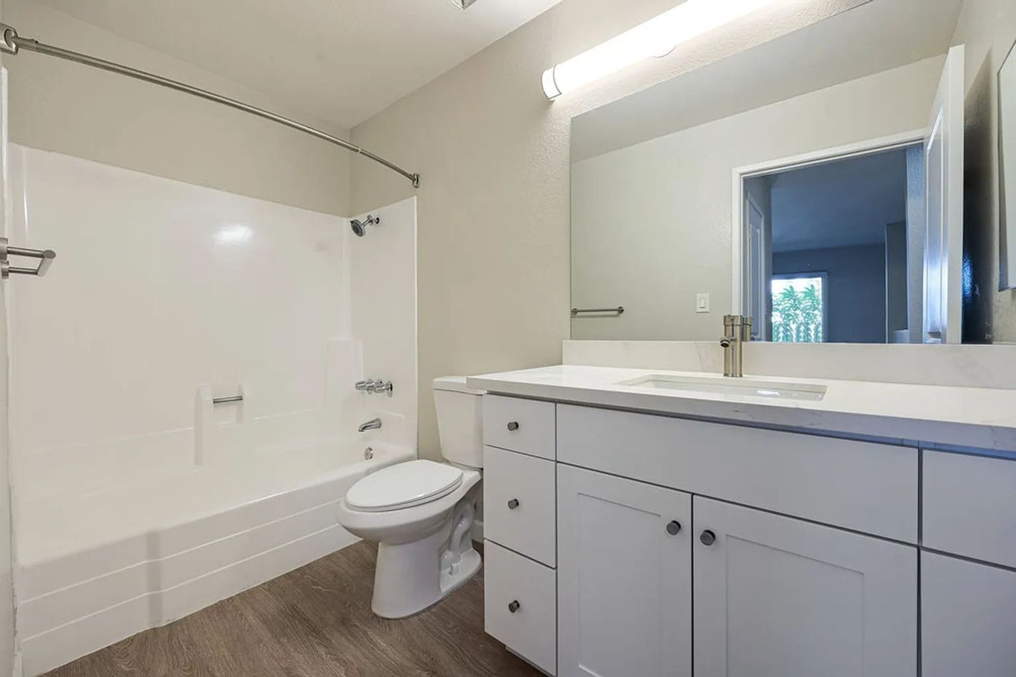 Bright white bathroom with bathtub, toilet, vanity sink, and large mirror on wood-look floor.