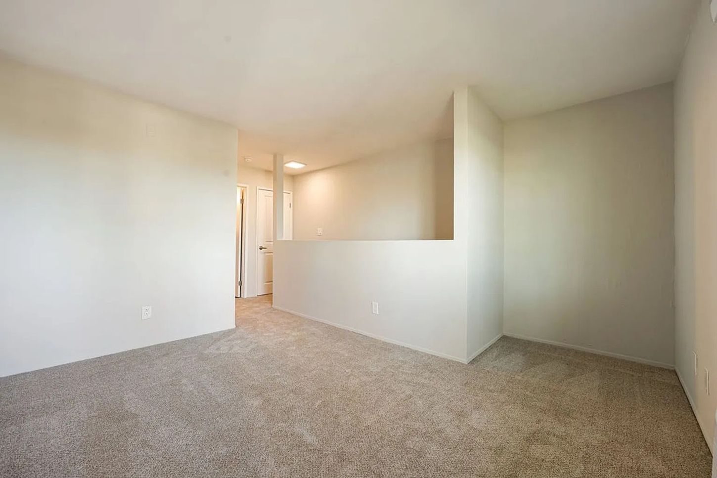Empty beige carpeted room with off-white walls and a partial half wall opening to a hallway