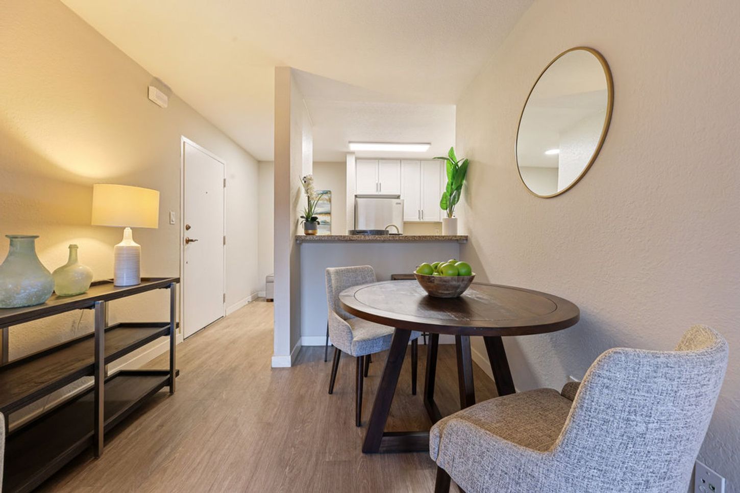 Small dining area with round table, chair, lamp, and view into a compact kitchen.