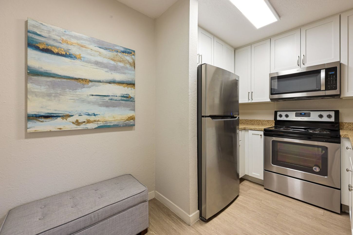 Small kitchen with stainless steel appliances, white cabinets, and a gray bench by abstract wall art.