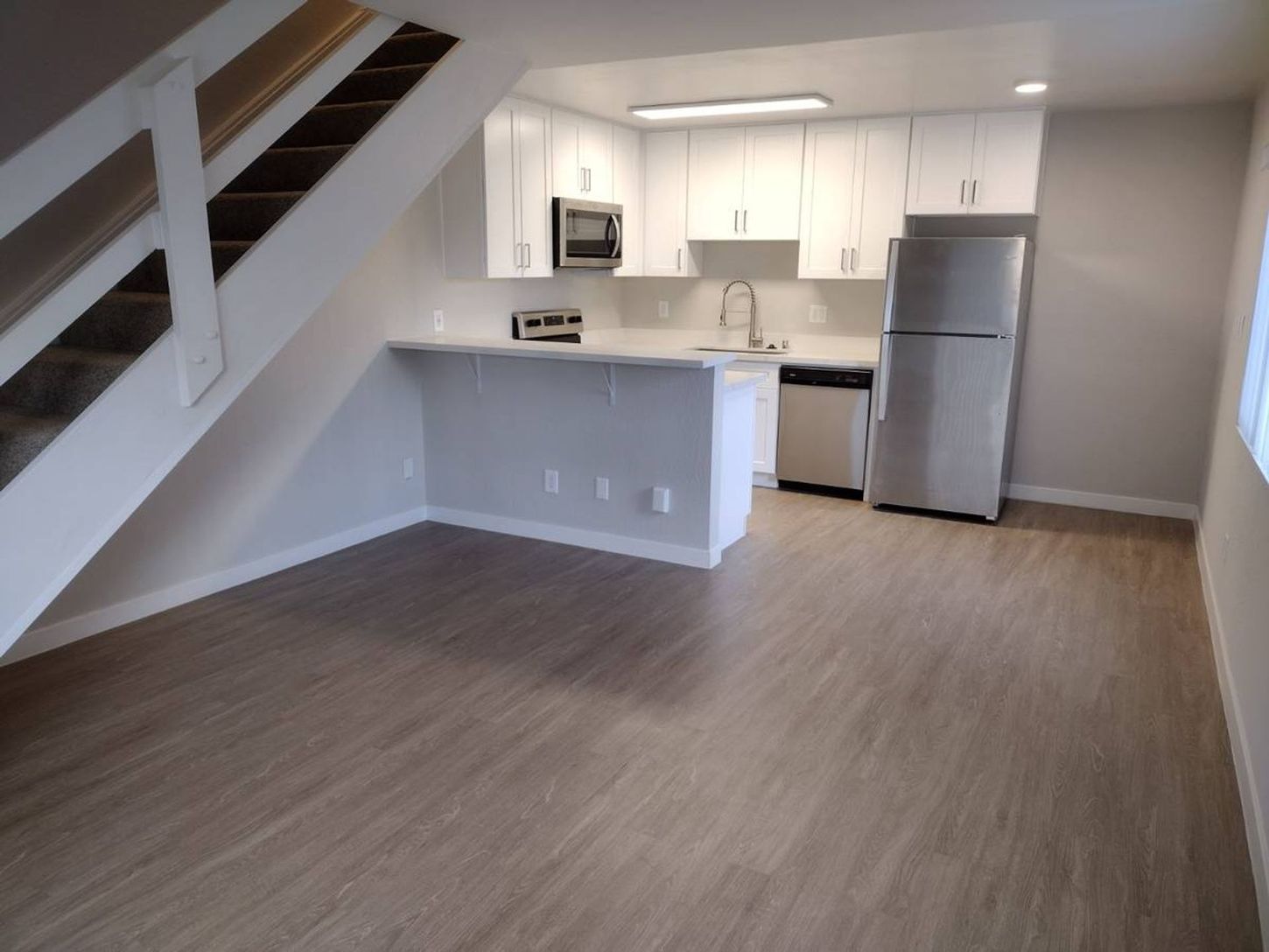 Empty open-plan kitchen and living area with white cabinets, stainless fridge, and staircase on the left.