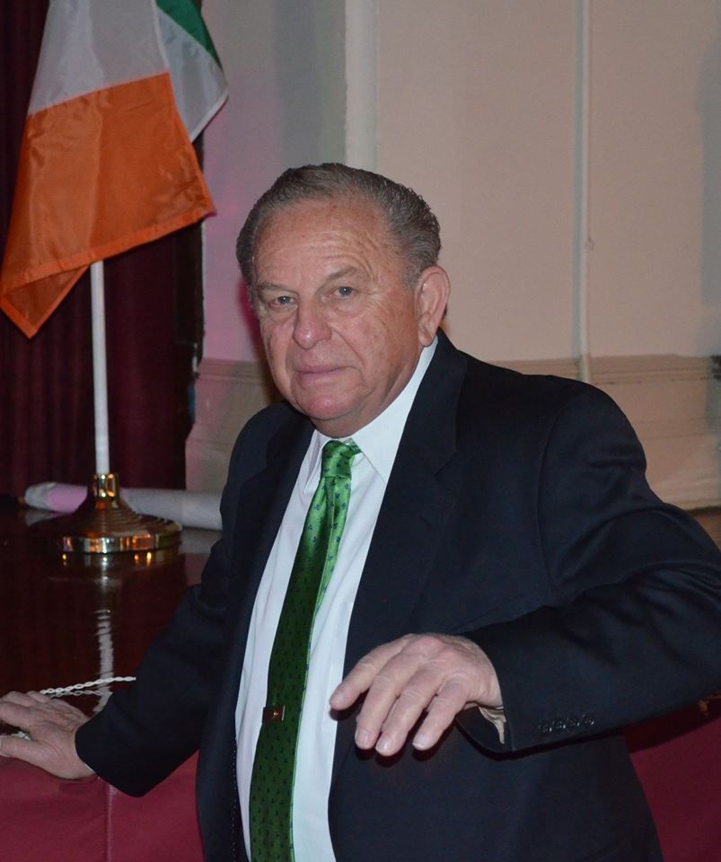 A man in a suit and tie stands in front of an irish flag