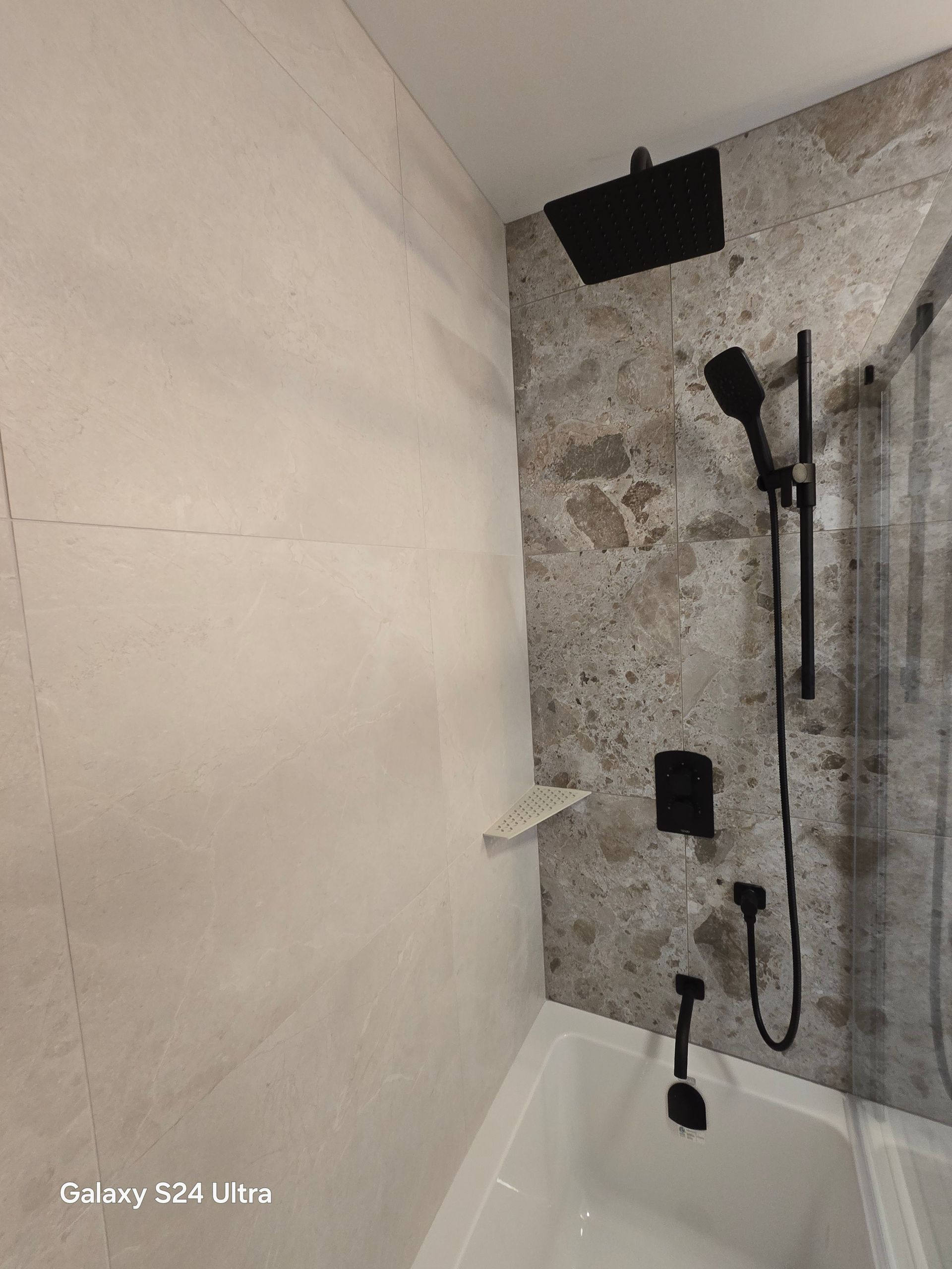 Robinetterie de douche noire dans une salle de bains carrelée de pierre et équipée d'une baignoire blanche.