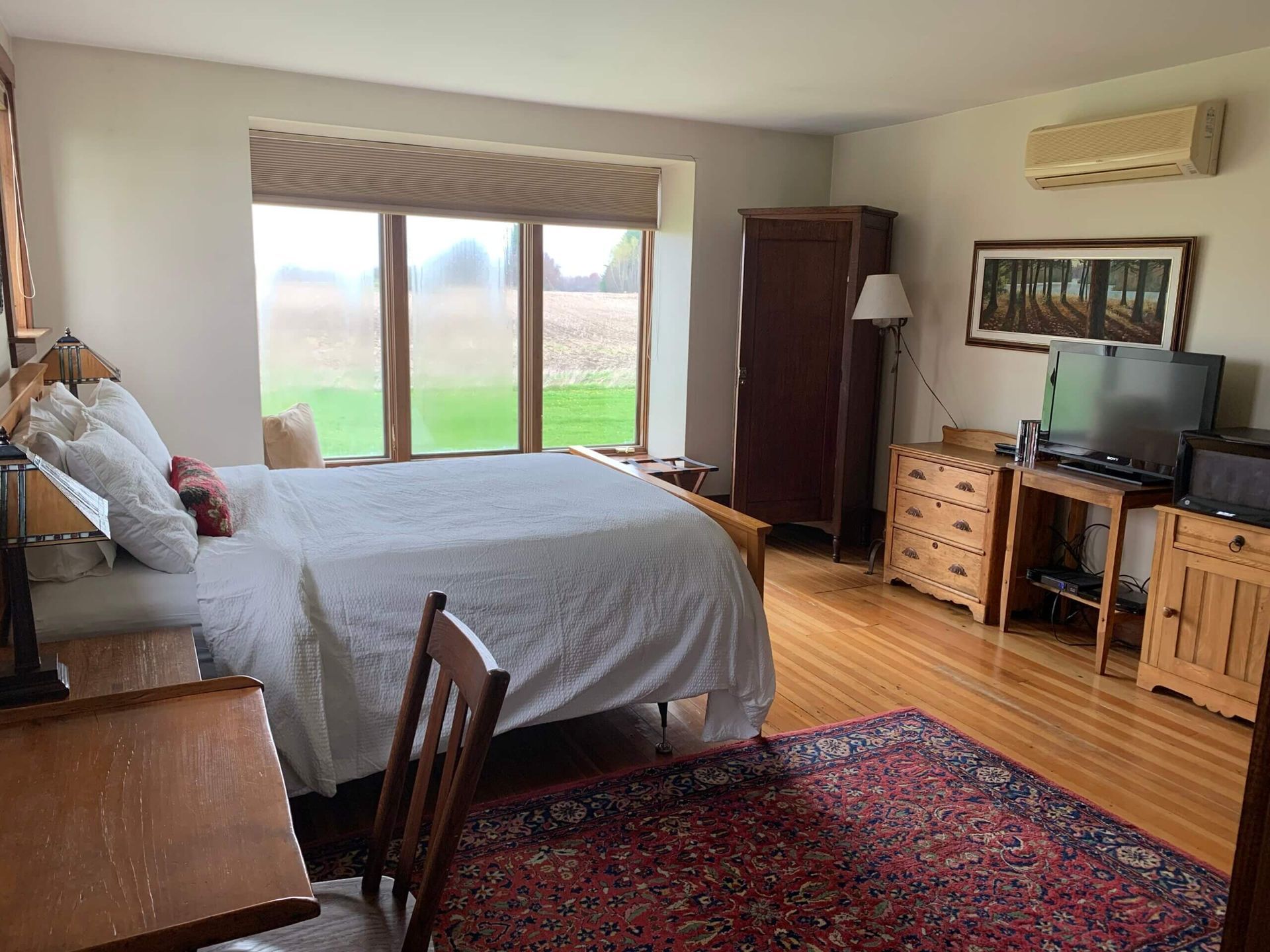 Bedroom with bed, furniture, large window, and rug. Hardwood floor and a view of grass.