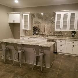 A modern kitchen with white cabinets, light marbled countertops, gray stools, and a mirrored backsplash.
