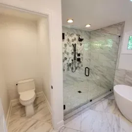 A bright, modern bathroom with a separate toilet nook, a glass-enclosed shower with floral tiling, and a white tub.