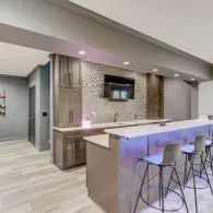 A modern basement wet bar with grey cabinetry, a chevron-patterned tiled wall, a mounted TV, and three bar stools.