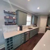 A modern kitchen with sage green cabinets, white countertops, open wooden shelving, and dark hardwood floors.