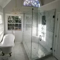 A bright bathroom featuring a white freestanding bathtub, a glass-enclosed shower, and an arched window with a chandelier.
