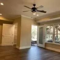 A bright, minimalist room featuring dark wood floors, beige walls, a ceiling fan, a white door, and a glass-paneled door.