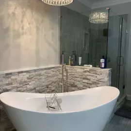 A modern bathroom featuring a white soaking tub, stacked stone wall, glass-enclosed shower, and decorative lighting.