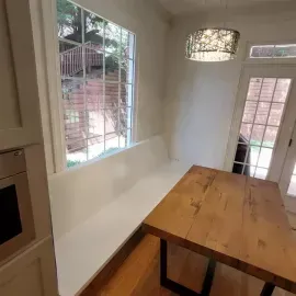 A built-in white bench seat sits next to a wooden dining table and a large window in a bright, modern kitchen.
