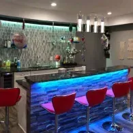 A modern home bar with red stools, stone-tiled counter with blue LED lighting, and four cylindrical pendant lights above.