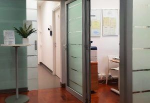 Moderno corridoio di uffici con porte a vetri, un piccolo tavolo da reception e uno spazio di lavoro open space visibile all’interno