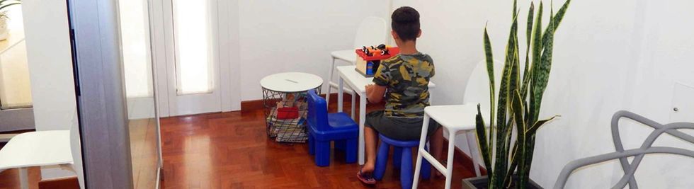 Bambino seduto a un tavolino in un corridoio, con sedie blu e una pianta alta in vaso nelle vicinanze