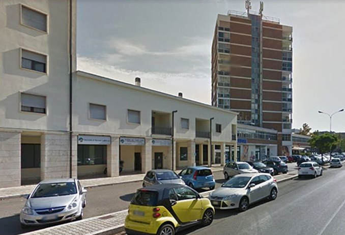 Strada con auto parcheggiate accanto a edifici bassi e a un alto condominio.
