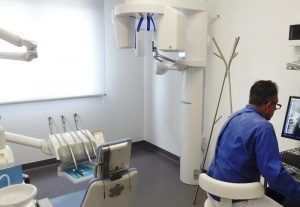 Studio dentistico con un paziente seduto sulla poltrona e il dentista al lavoro davanti a un monitor, con un apparecchio radiografico sopra la testa.