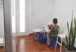 Bambino seduto a un tavolino bianco in una stanza luminosa, con sgabelli blu e piante in vaso