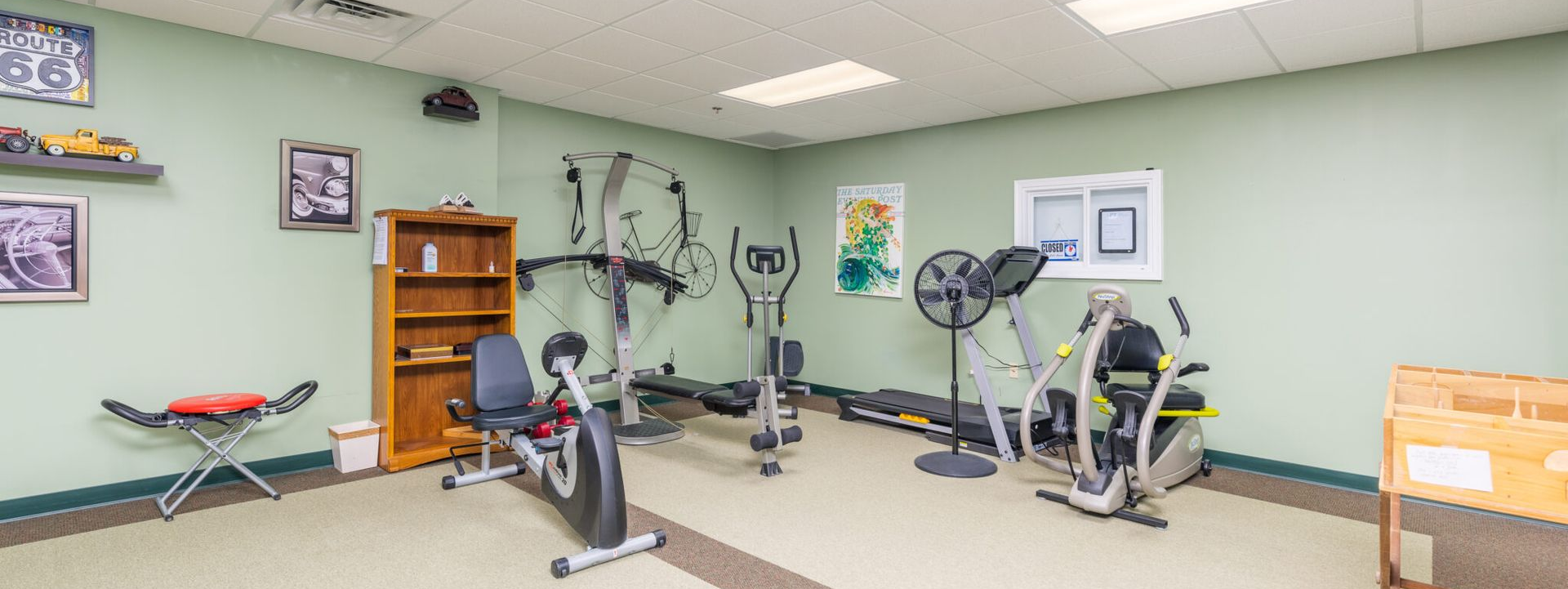 A small gym with exercise machines and weights. Light green walls, wooden shelves, and a beige and brown floor.