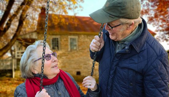Elderly couple on a swing set, smiling at each other in front of a house, surrounded by fall foliage.