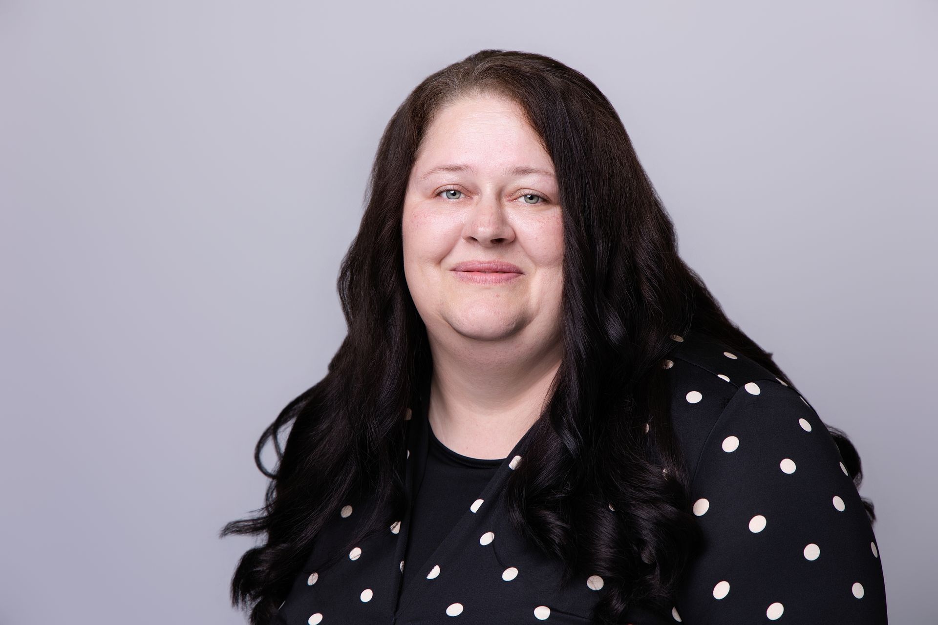 Person with long dark hair smiling against a gray background, wearing a black polka-dot top.