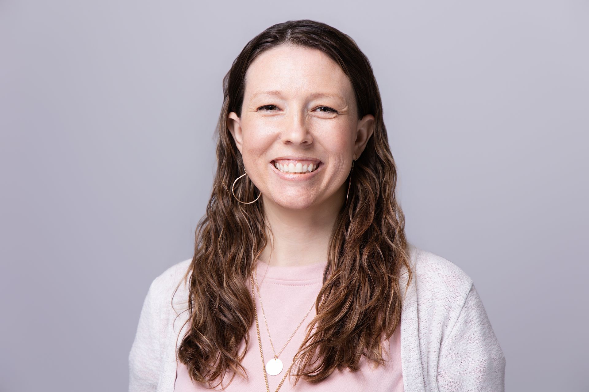 Smiling woman with long brown hair in a white cardigan and pink top against a gray background