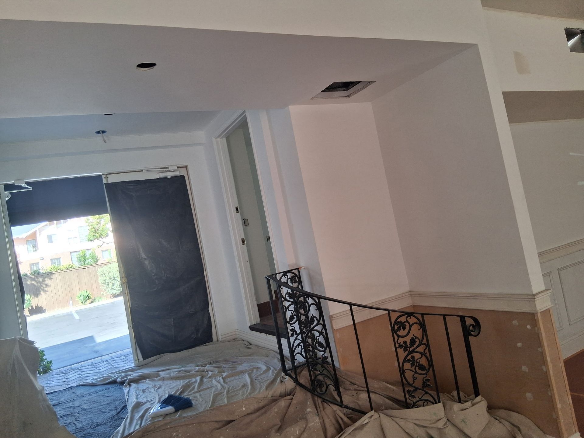 An interior space undergoing renovation, featuring white walls, a black iron stair railing, and protective drop cloths.