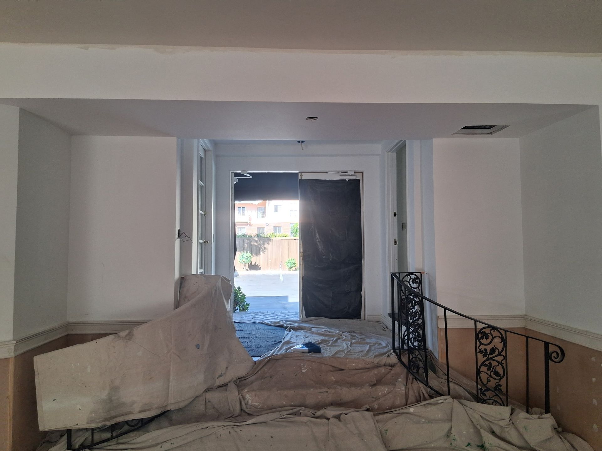 An interior view of a room under renovation, showing a doorway leading outside, covered in drop cloths and dust.