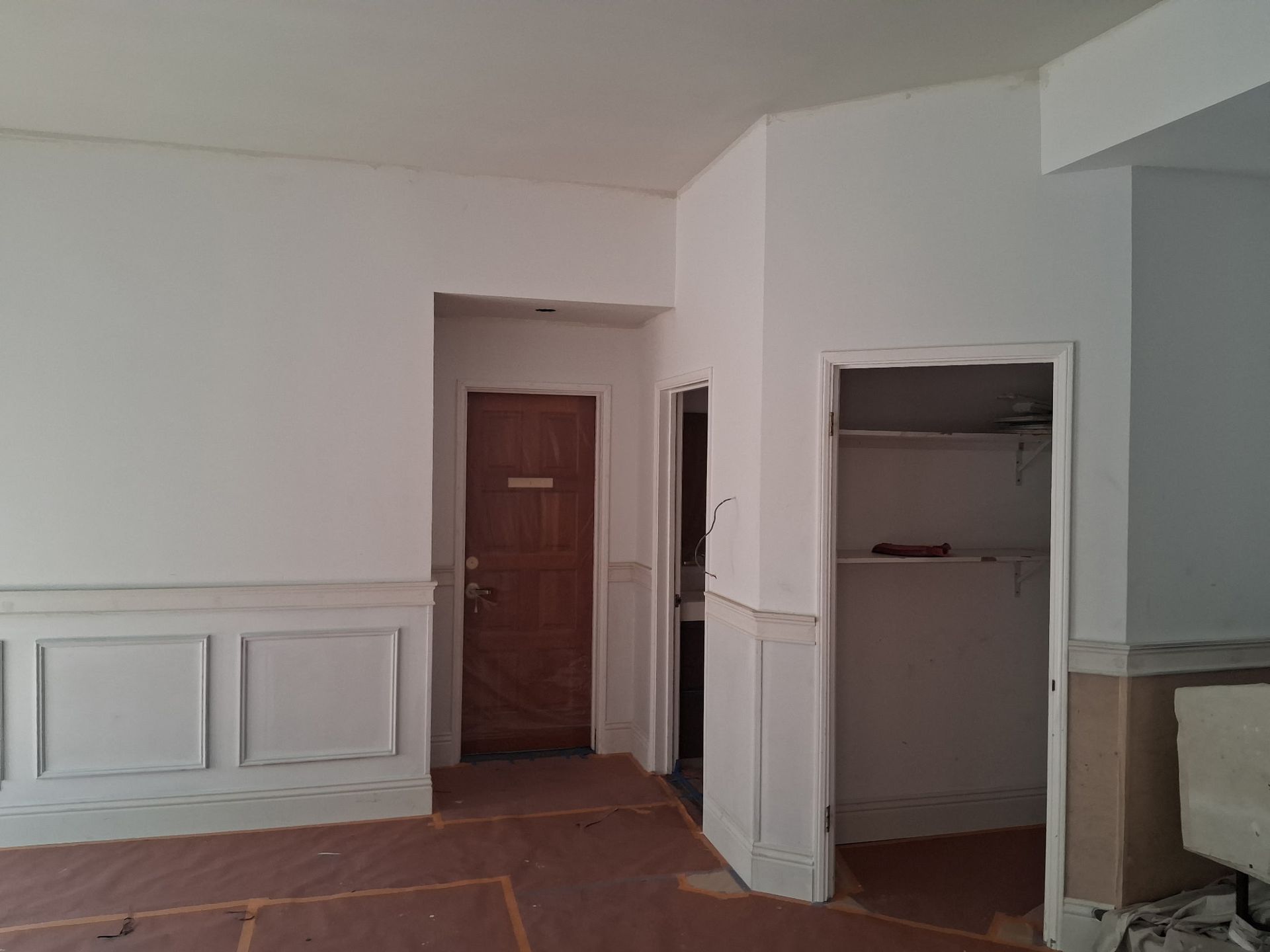An interior view of a room undergoing renovation with white walls, wainscoting, a wooden door, and an open closet.