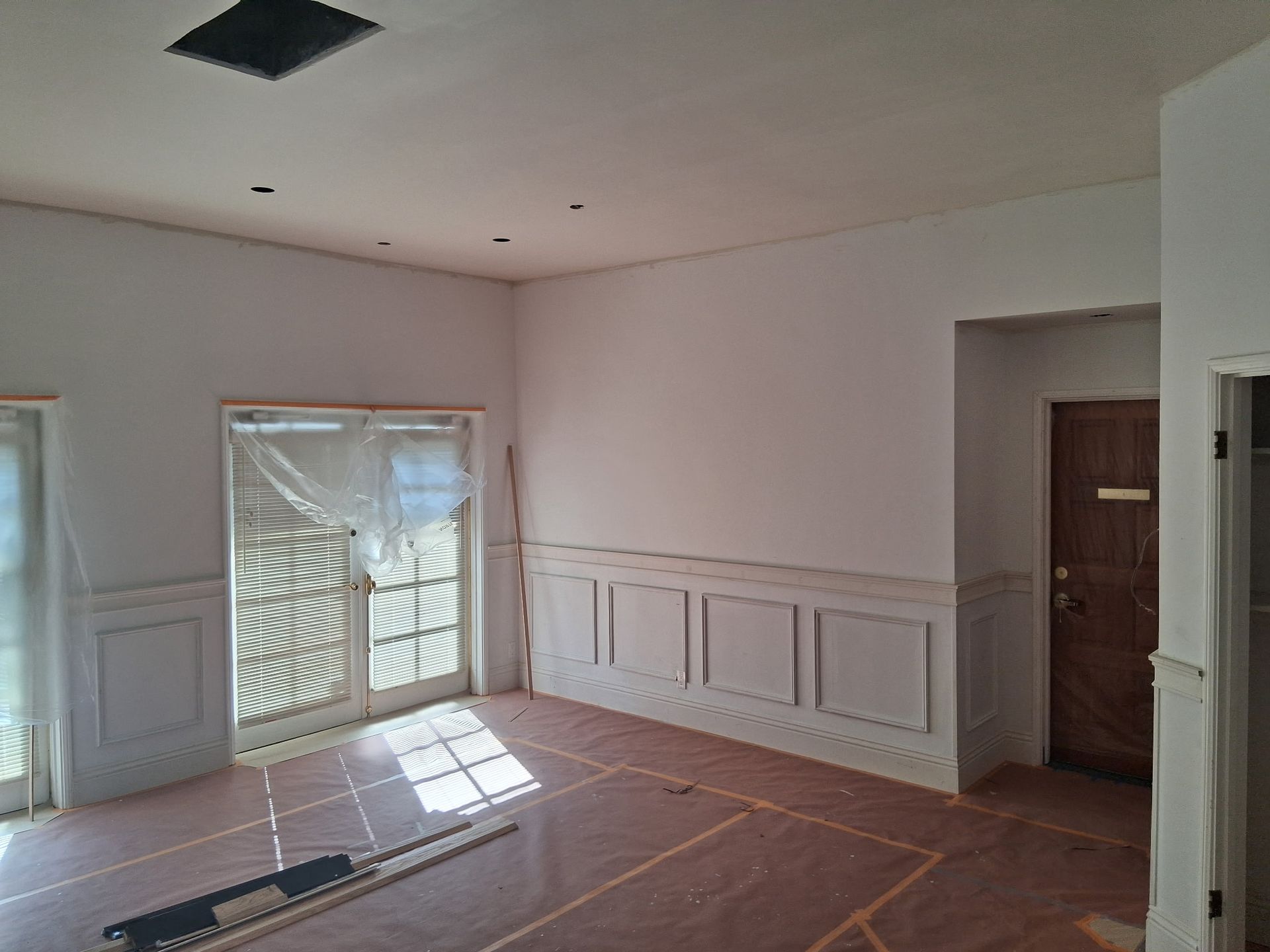 A room under renovation with white walls, wainscoting, glass doors covered in plastic, and a brown wooden door.