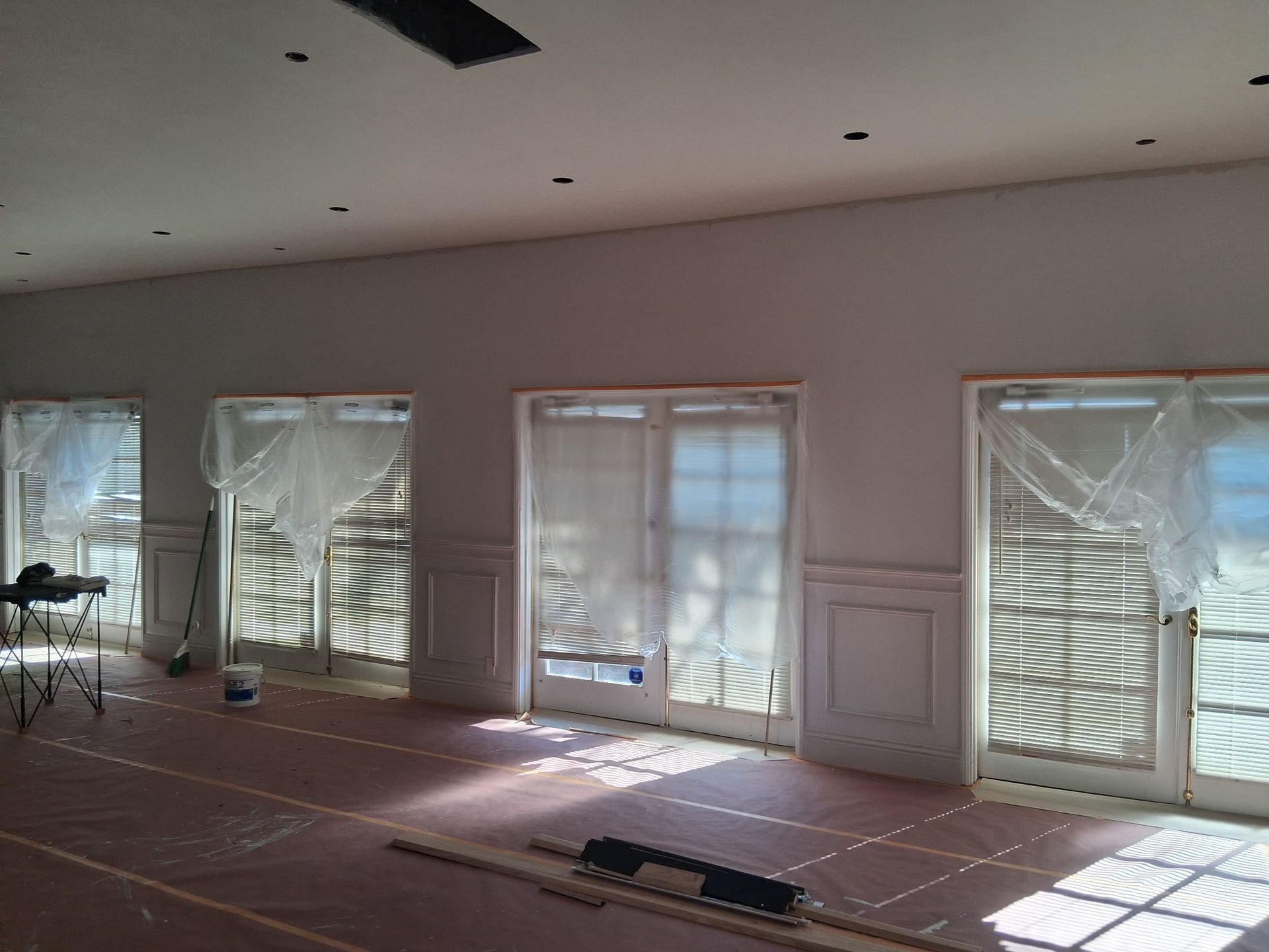 A large, bright, freshly painted white room with paneled walls, four glass doors covered in plastic, and floor protection.