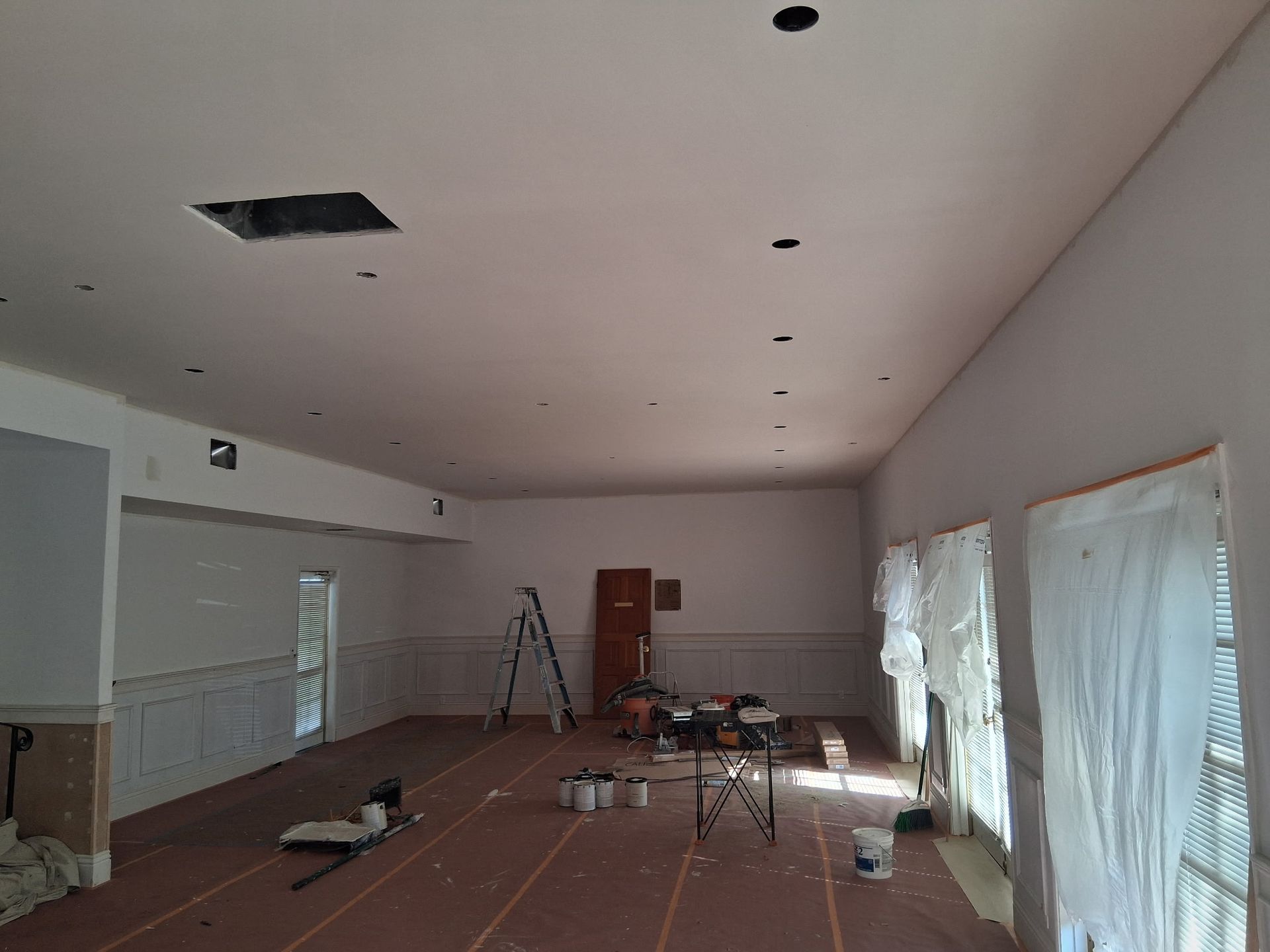 Interior of a room under construction with white walls, a freshly plastered ceiling, a ladder, and drop cloths on windows.