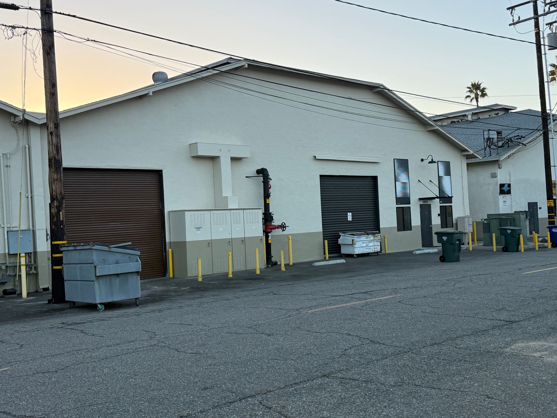 A tan industrial warehouse exterior with two large dark roll-up doors, a white cross, and several trash bins out front.