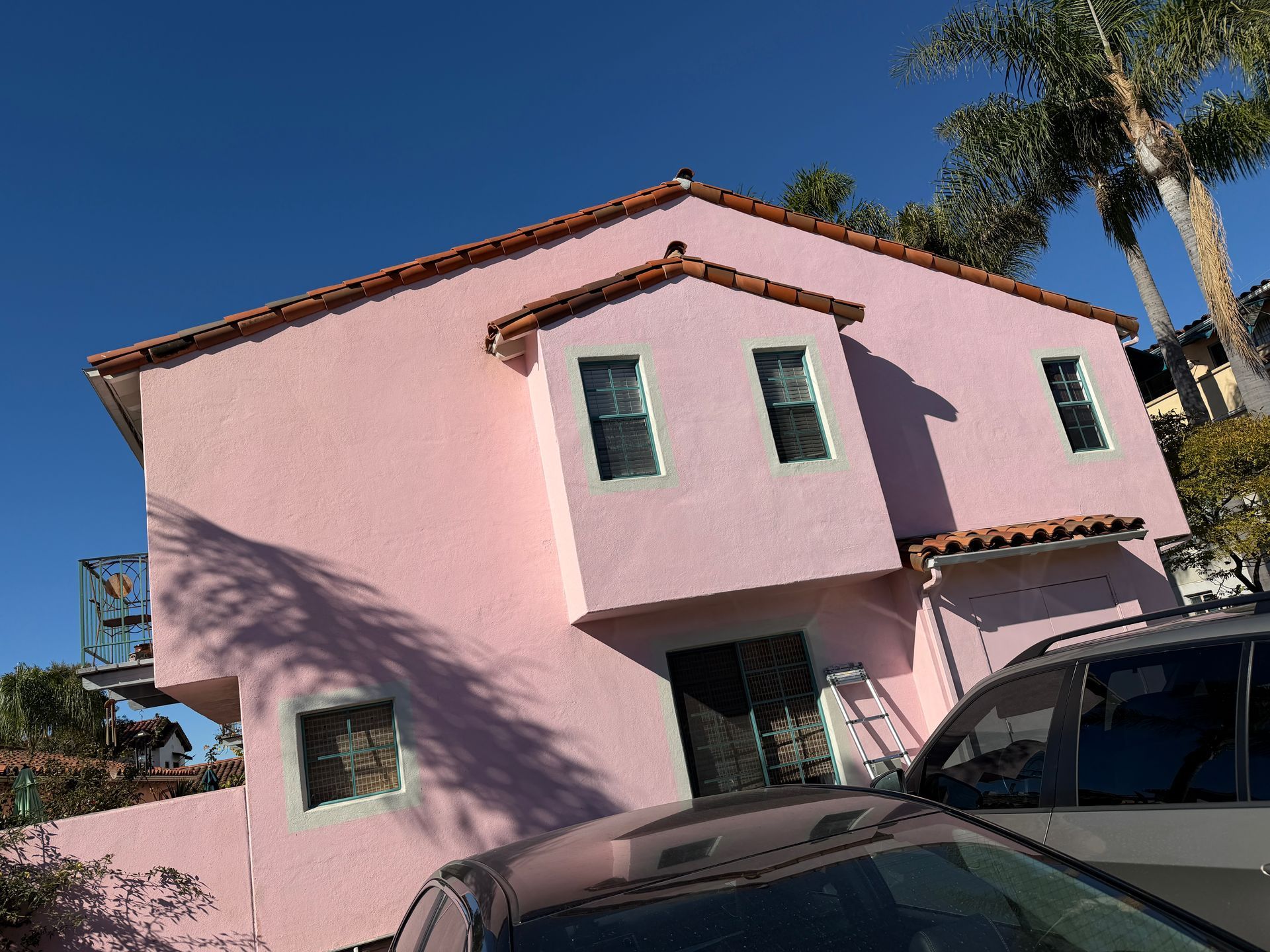A two-story pink stucco house with a tiled roof under a clear blue sky, partially obscured by a car in the foreground.