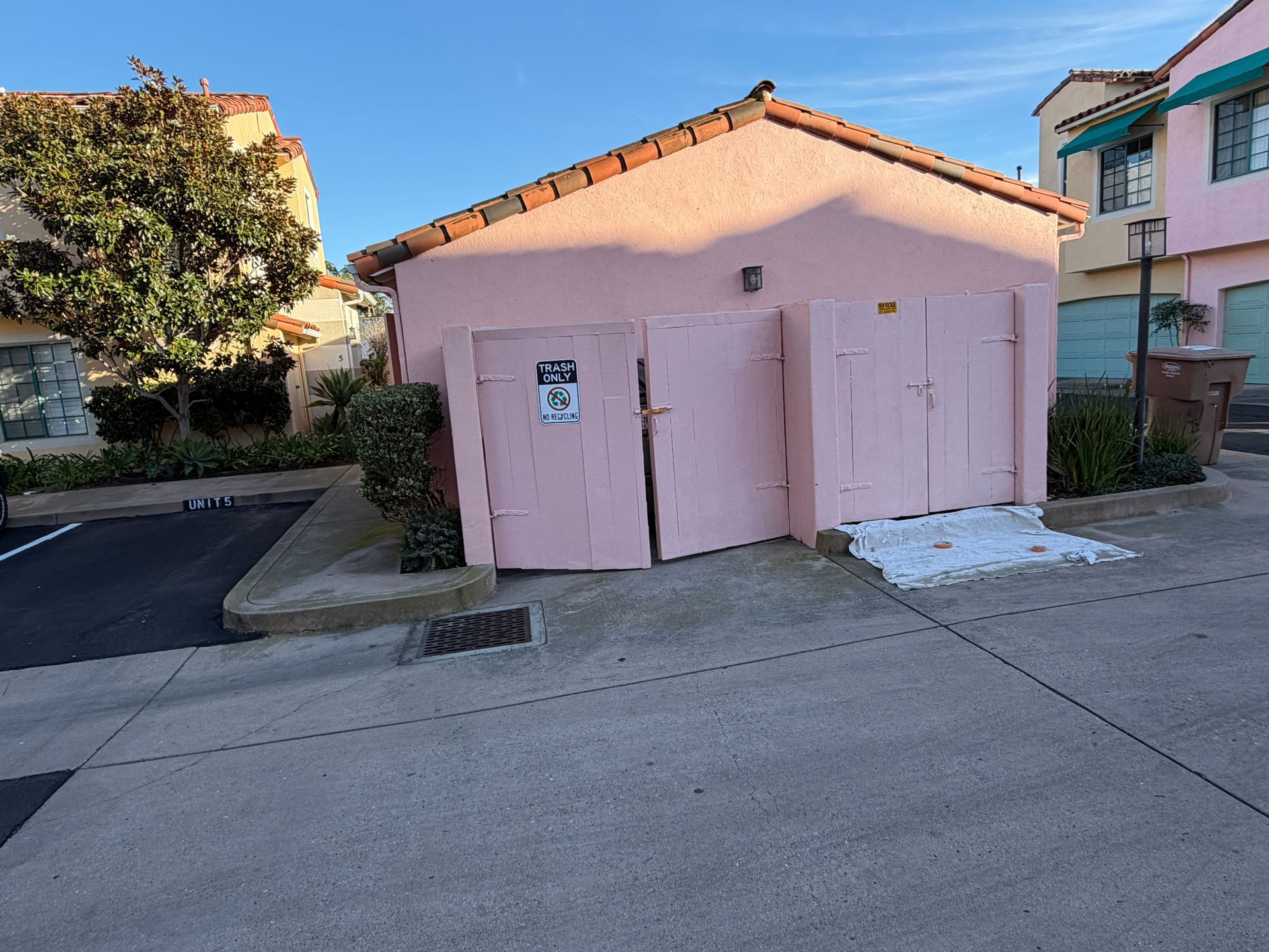 A small, pink, shed-like structure with double doors and a tiled roof, located in a concrete parking area under blue sky.