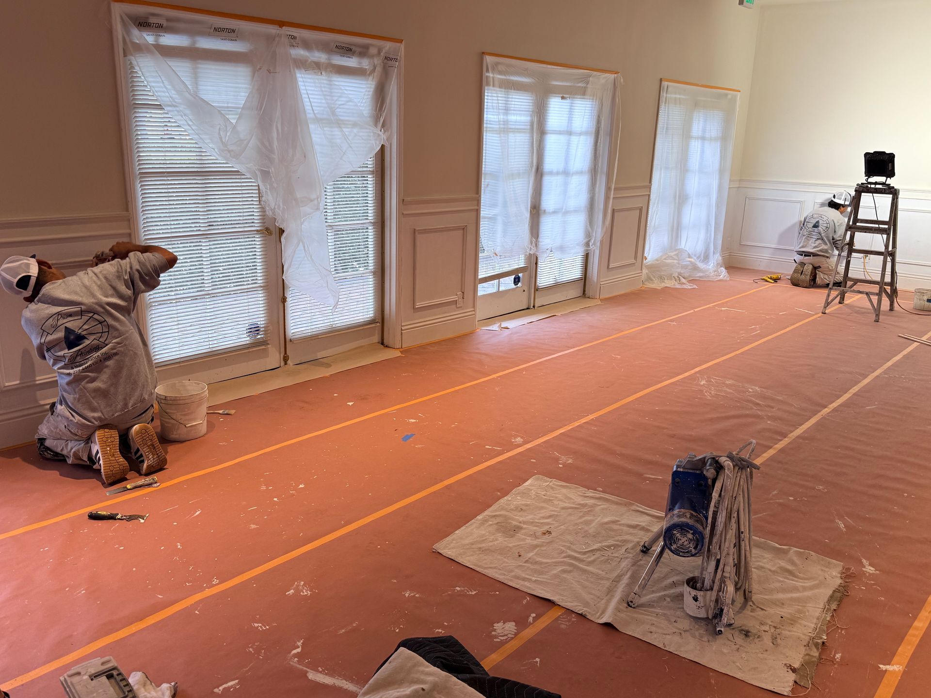 Two painters in white coveralls work inside a room with plastic-covered windows and orange floor protection.