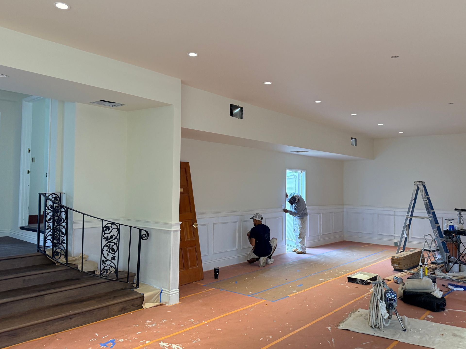 Two people work on installing wainscoting on a wall in a room with a staircase, covered floors, and recessed lighting.