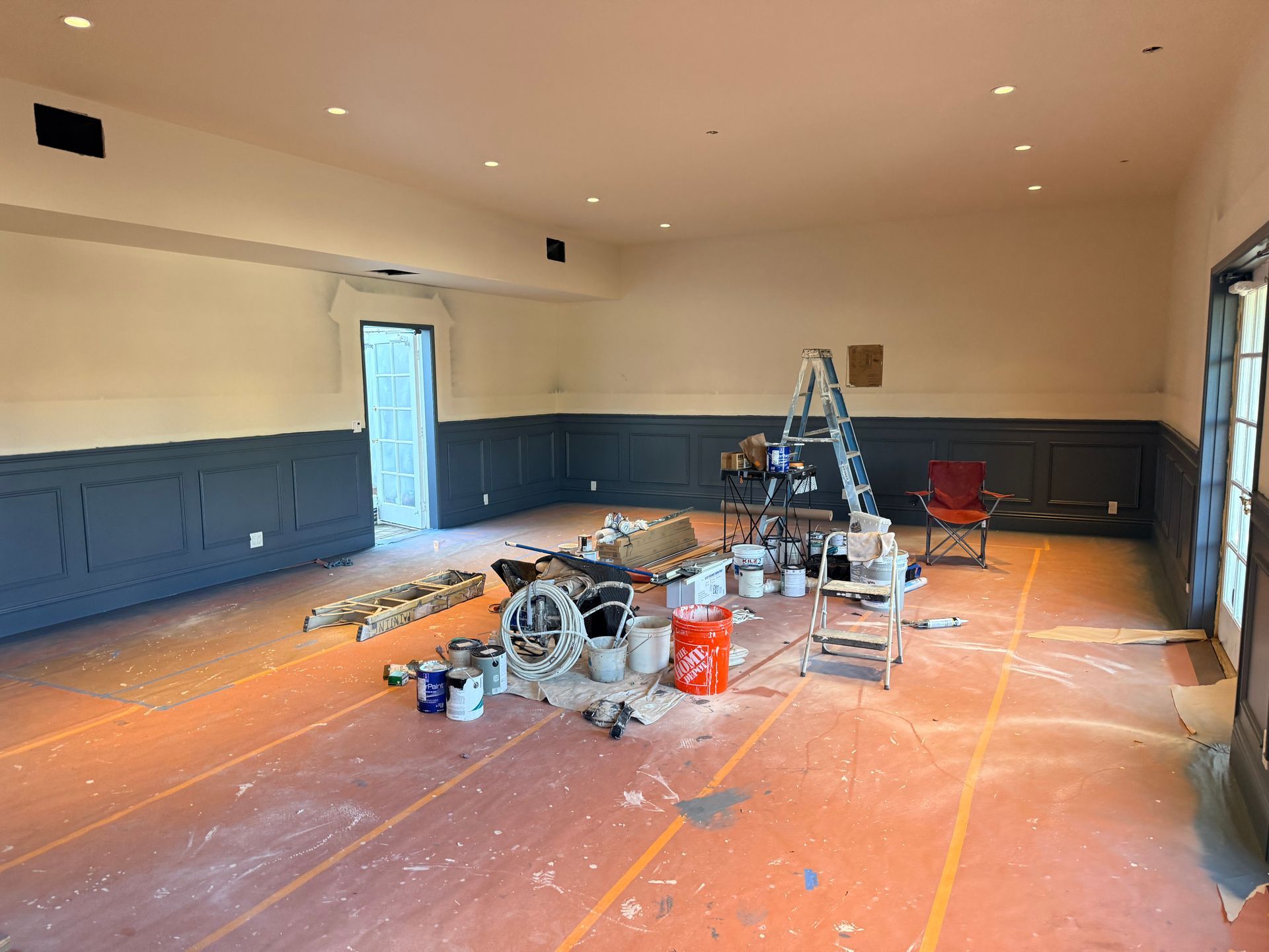 A room under renovation with dark grey wainscoting, white walls, and painting equipment scattered across floor paper.