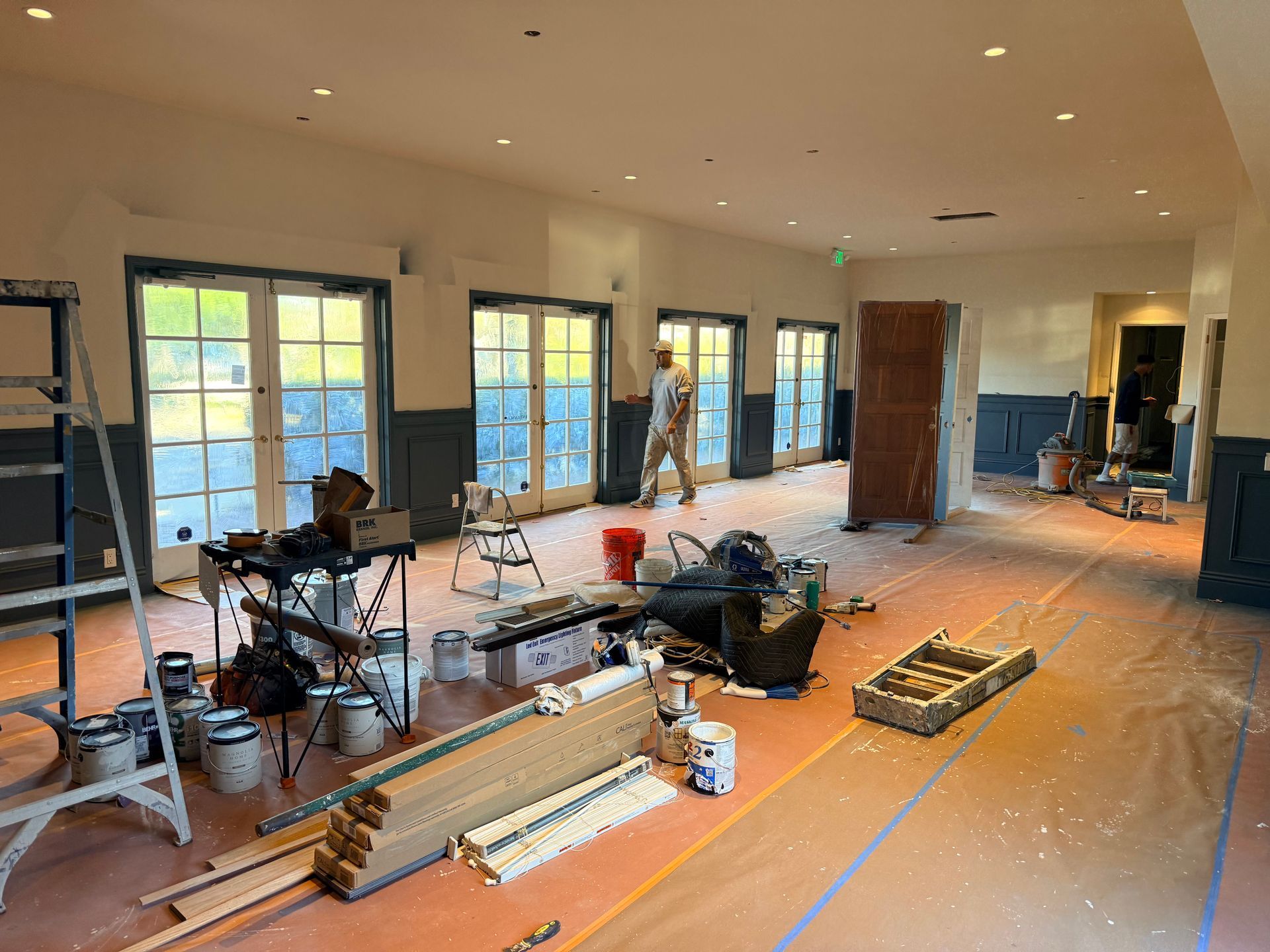 A person works in a large room under renovation, with drop cloths on the floor, construction materials, and paint supplies.