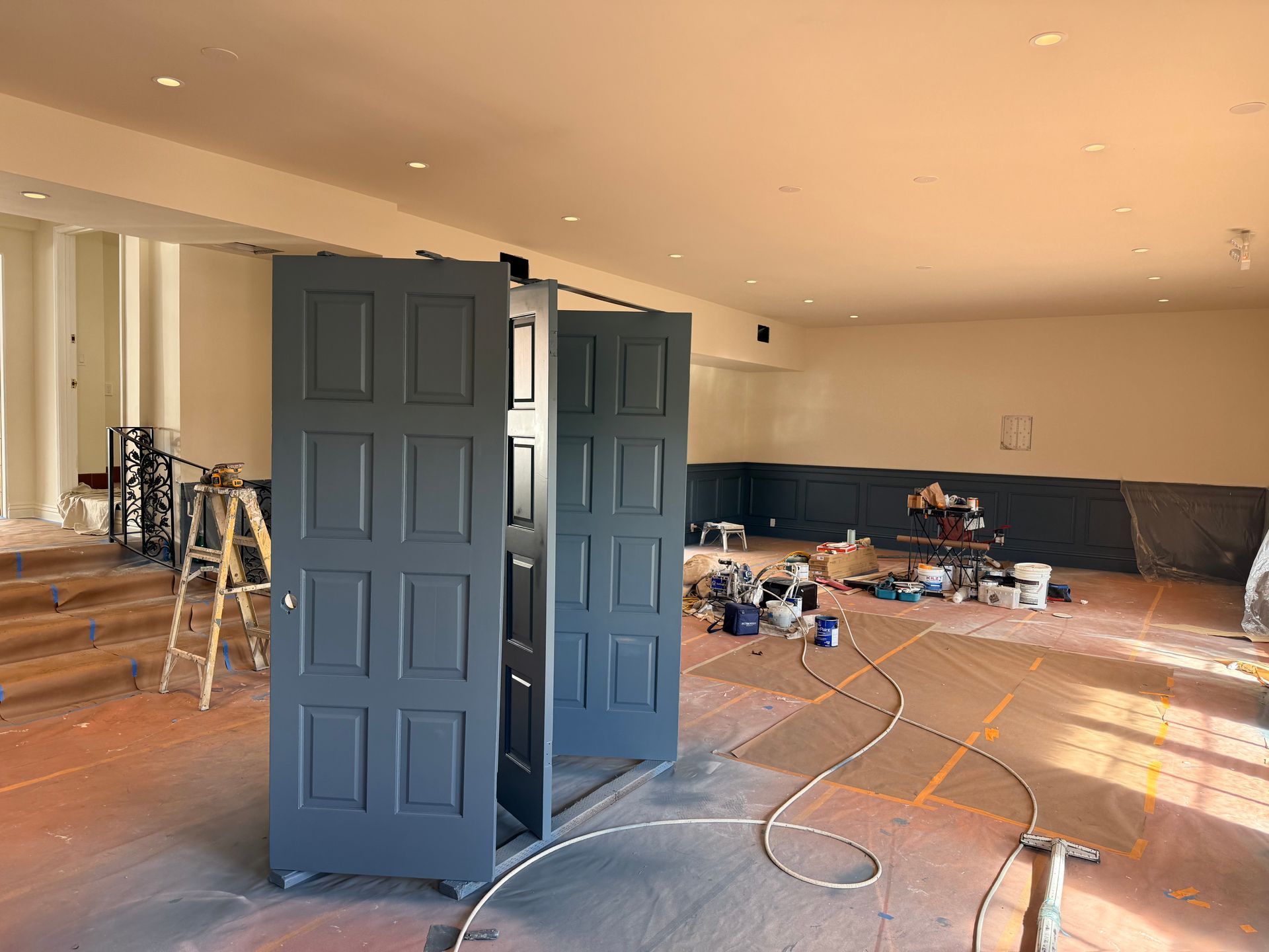 A dimly lit room under construction with dark gray painted doors standing upright and paint supplies scattered on the floor.