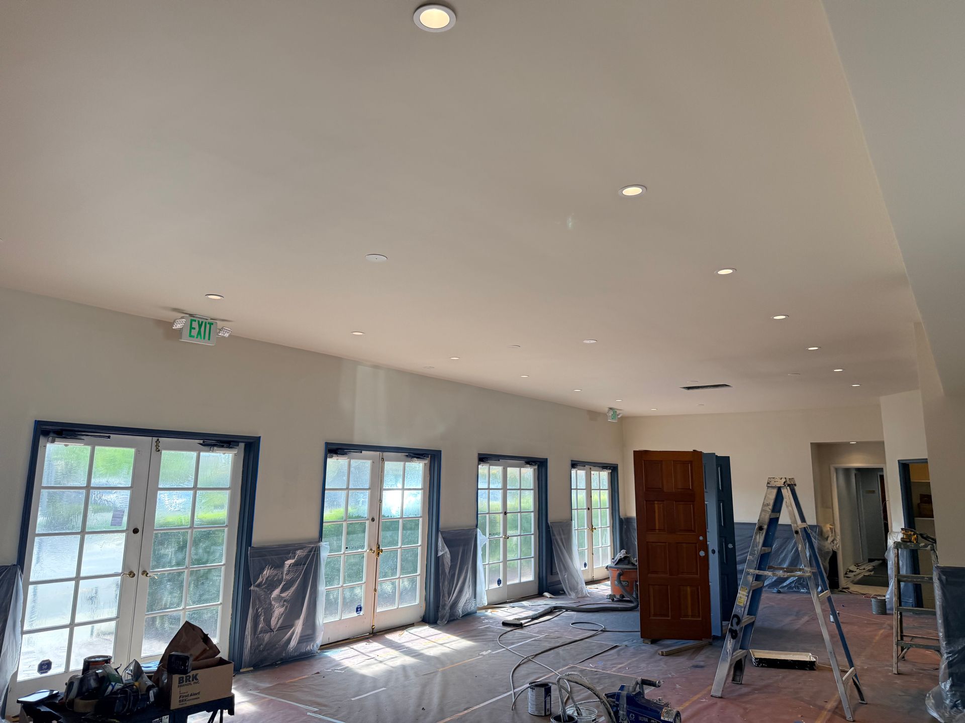 Interior of a room under renovation with drop cloths on windows, a wooden door, and a metal ladder on a wood floor.