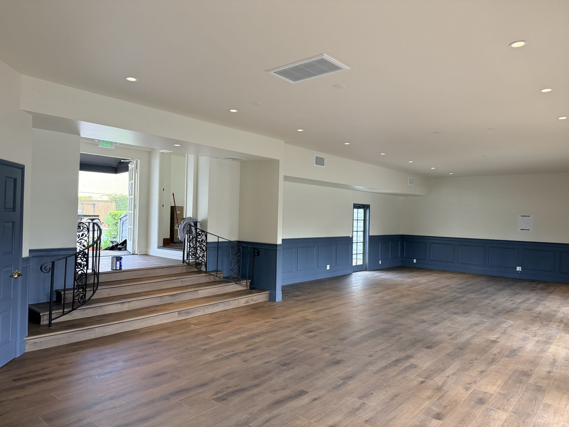 A spacious room with light hardwood floors, white walls, and a dark blue wainscoting trim, featuring a short set of stairs.