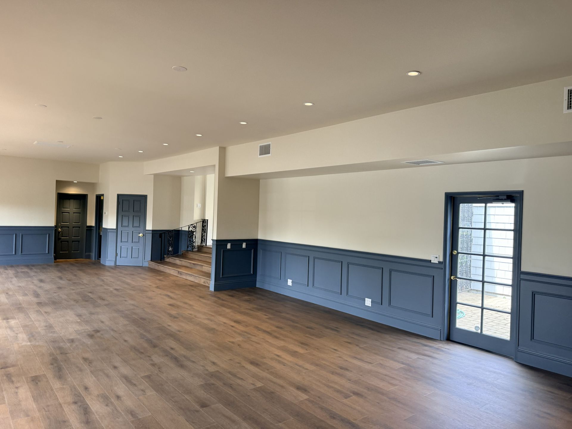 A spacious room with dark blue wainscoting, light-colored walls, wood flooring, and a glass-paned door.