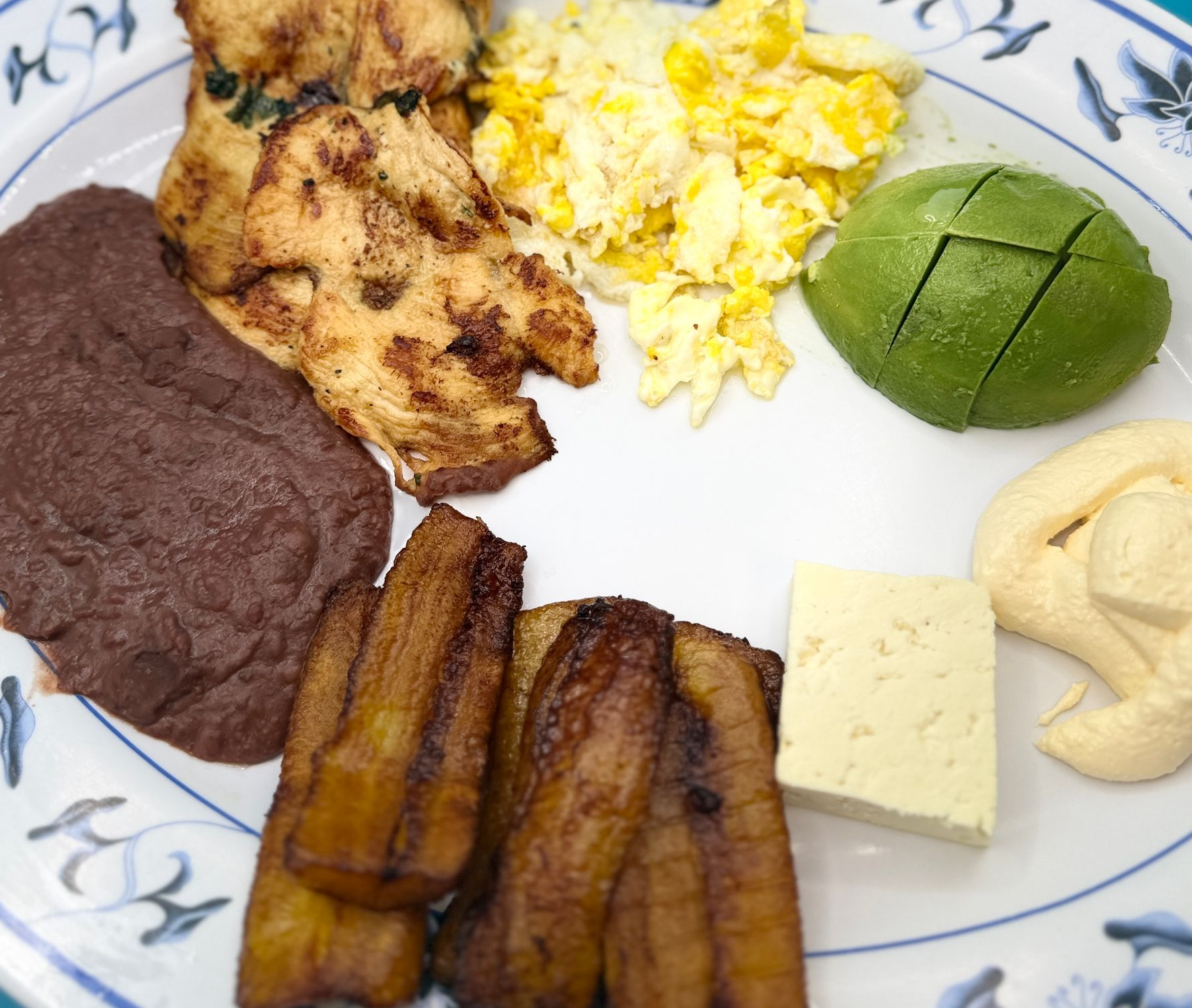 Plate of assorted foods: refried beans, grilled chicken, scrambled eggs, avocado, plantain slices, cheese, and cashews