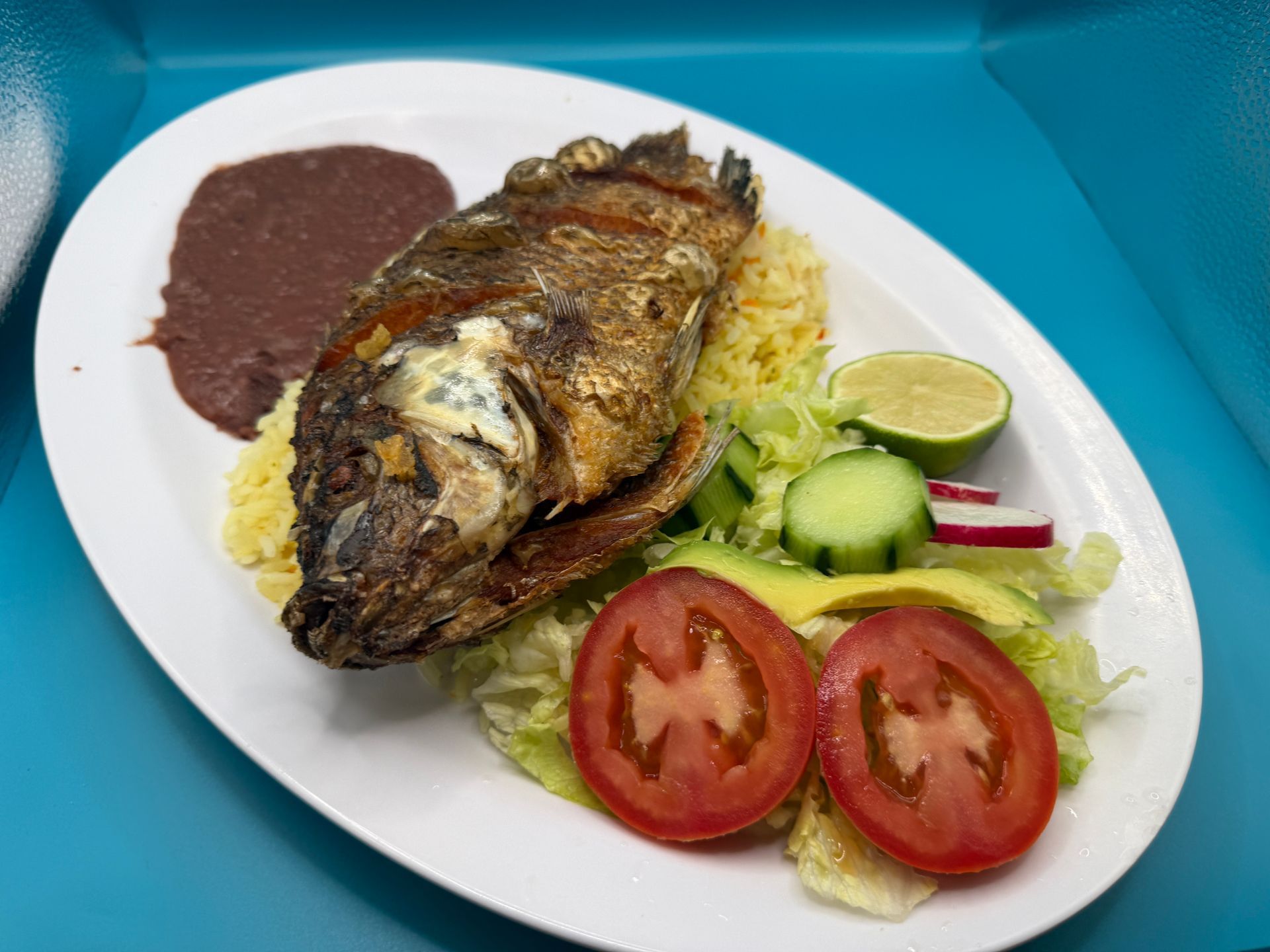 Grilled whole fish with rice, beans, salad, and lime on a white plate with blue tablecloth