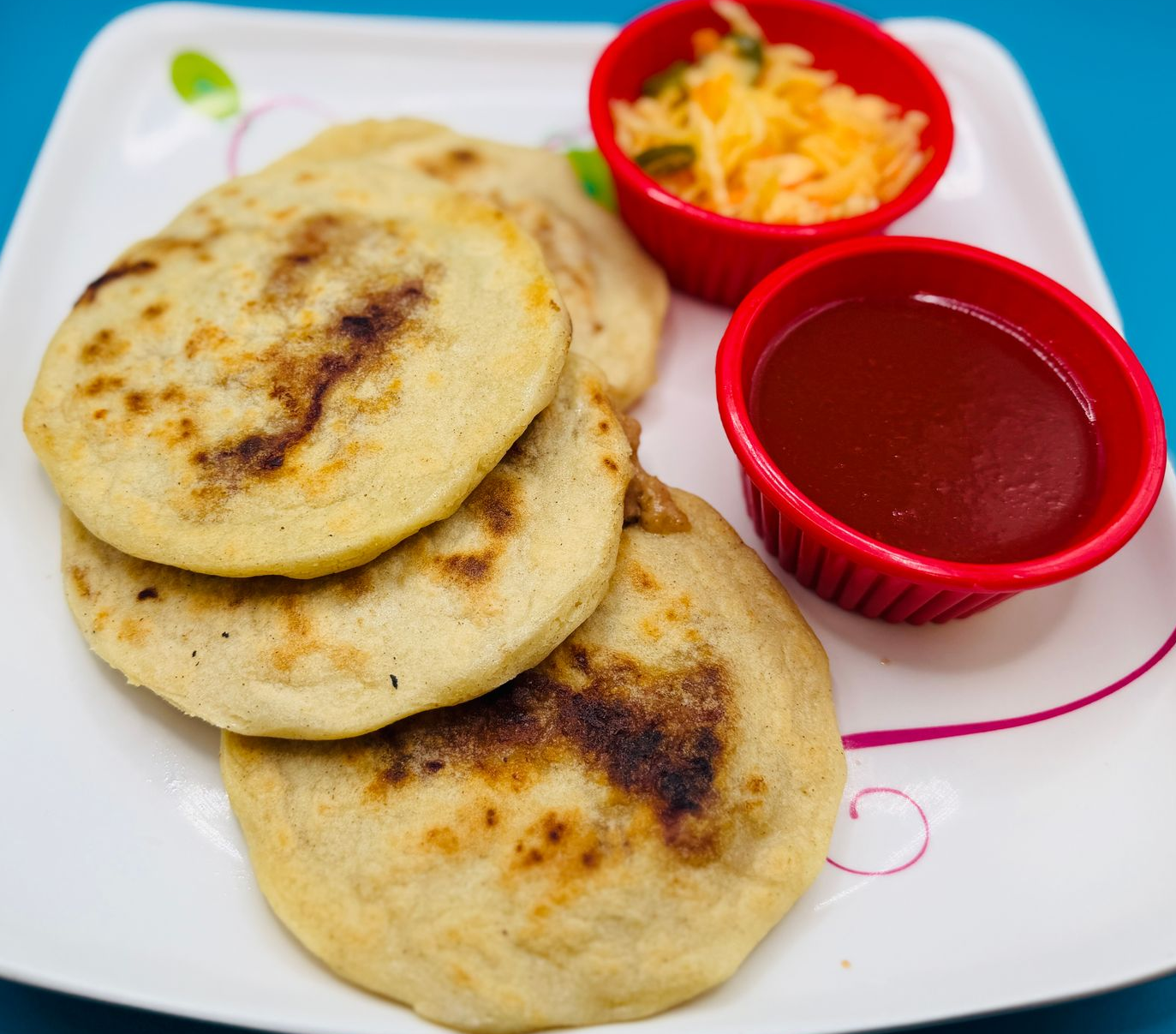 Three arepas on a white plate with red cups of sauce and shredded cheese.