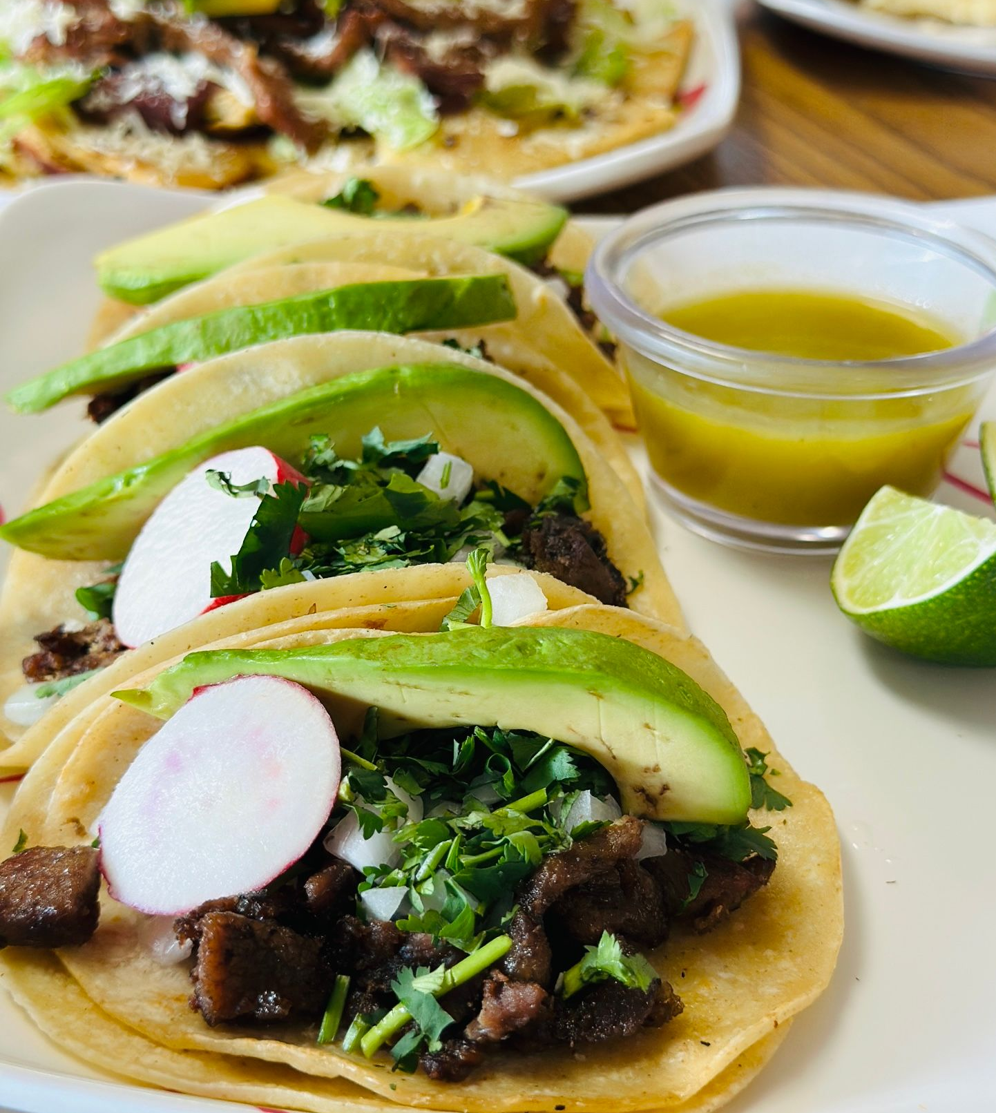 Tacos topped with avocado and radish, with salsa and lime on a table