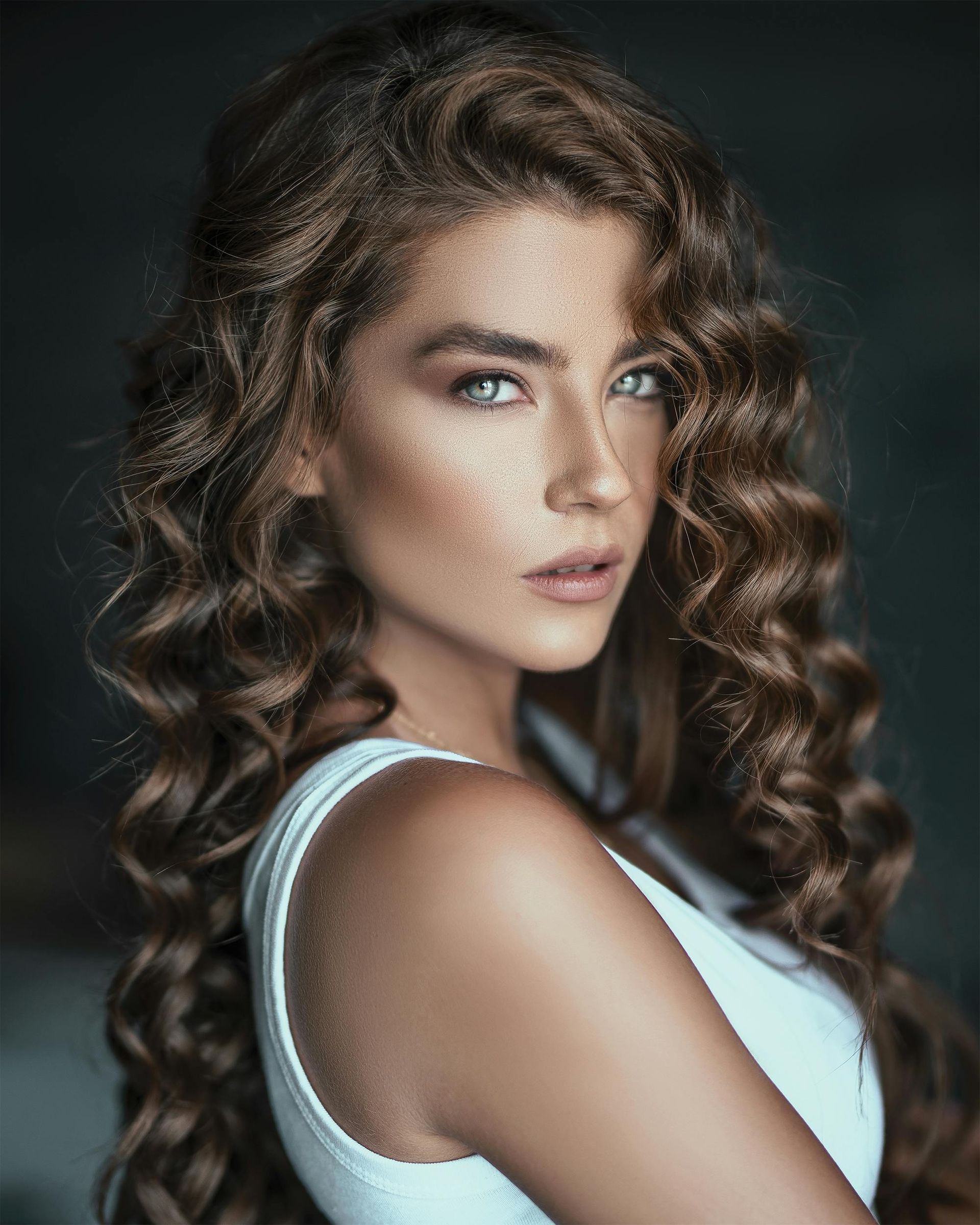 A woman with long curly hair is wearing a white tank top and looking at the camera