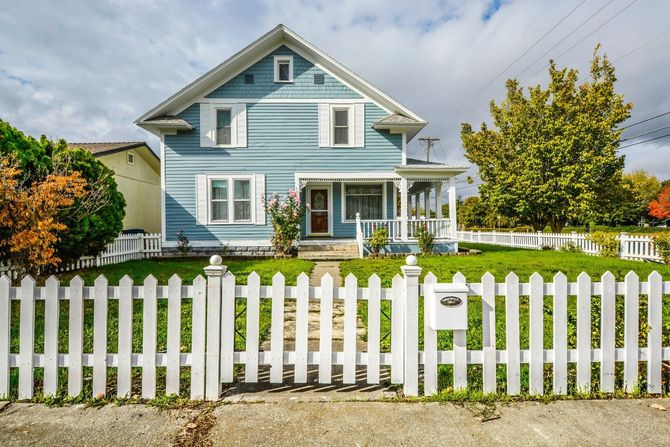 Blue house with white trim behind a white picket fence and green lawn under a cloudy sky